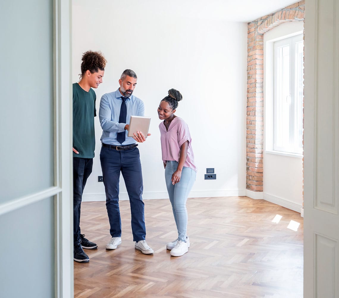 A realtor and two clients review a tablet while standing in an empty room, exploring options for house hunting.